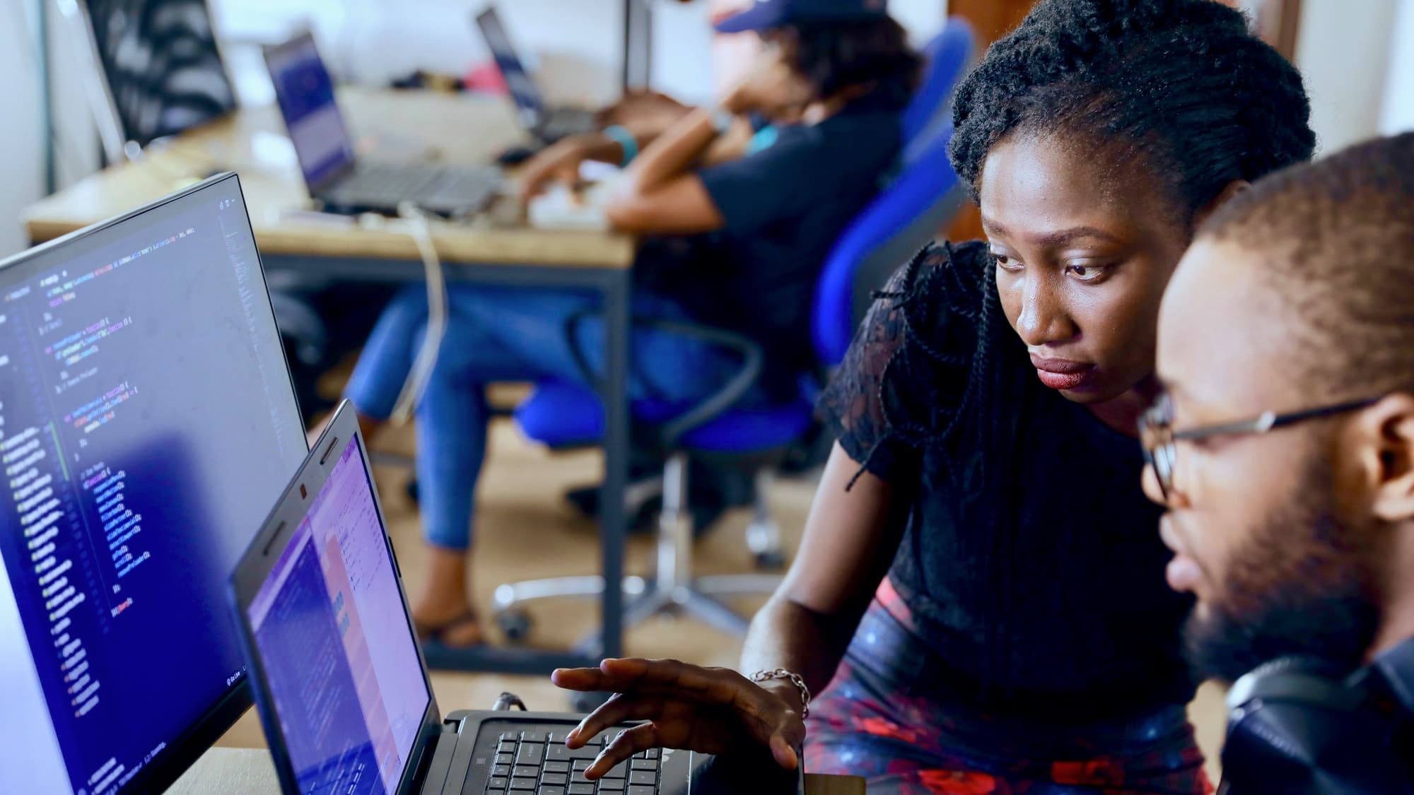 Two colleagues working through code together on a laptop in a busy office